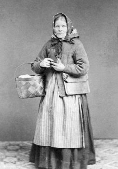 Ett foto från 1869 av en medelålders kvinna i huckle, lång kjol, förkläde, tröja, en platt postväska över axeln och en spånkorg över höger armbåge, där något vitt, troligen tyg, skymtar